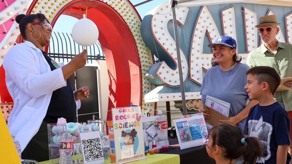 A family enjoying an educational presentation in front of unlit neon signs arranged outdoors in daylight