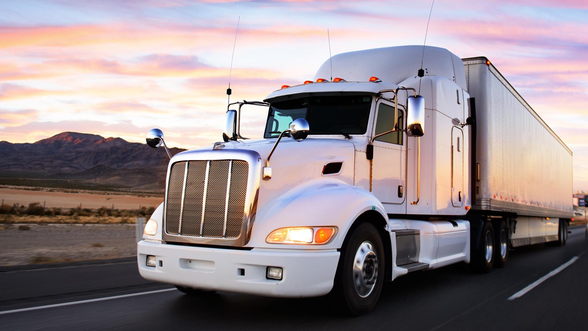 A large white truck drives down a desert freeway as the sun is setting, casting a pink and purple glow on the clouds in the darkening sky