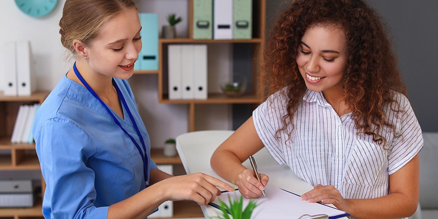 A medical assistant wearing blue scrubs stands behind a counter pointing at paperwork that a patient with curly hair and wearing a white and blue striped shirt is filling out with a pen
