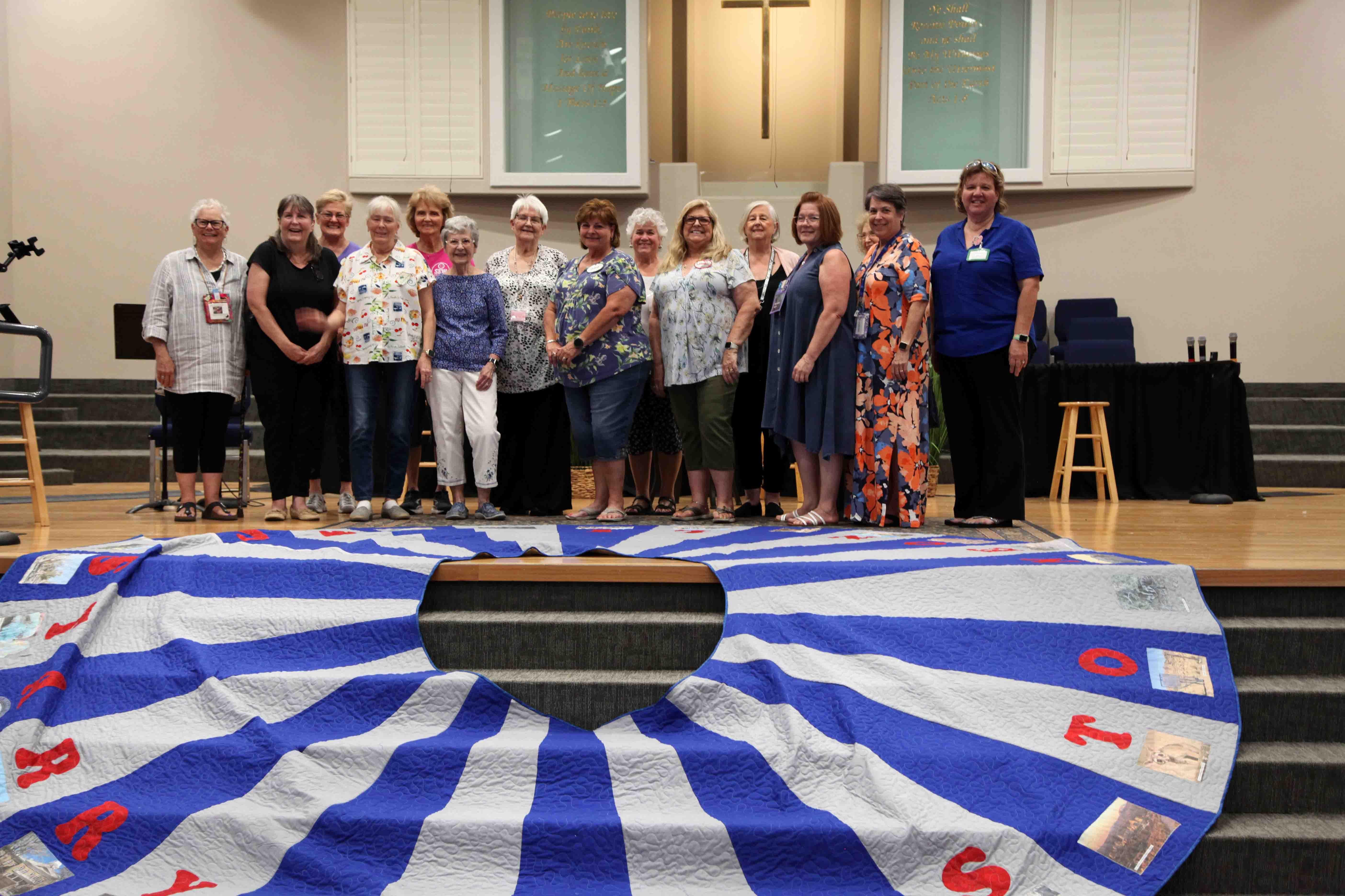 A line of volunteers stand on the altar of a church behind a large quilted Christmas tree skirt splayed out on stairs