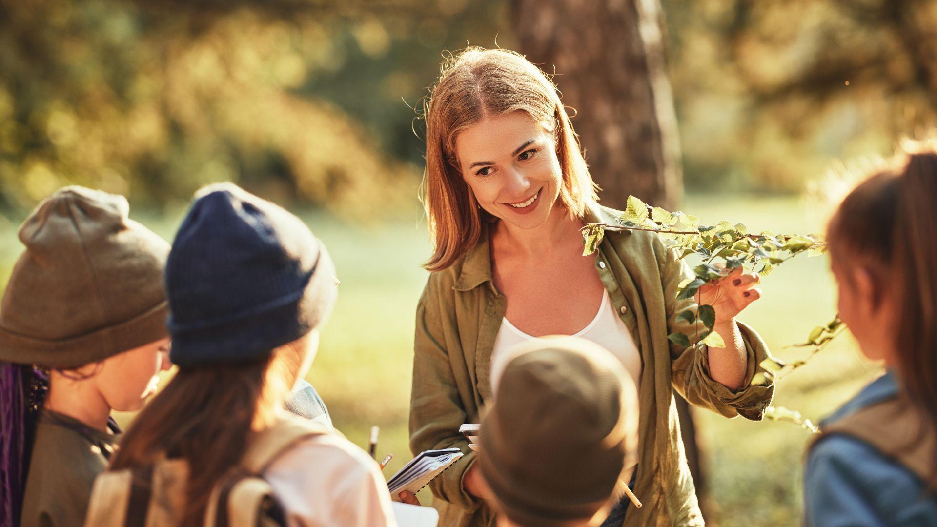 A Girl Scout Guide hold some branches while four girls gather around her outside.