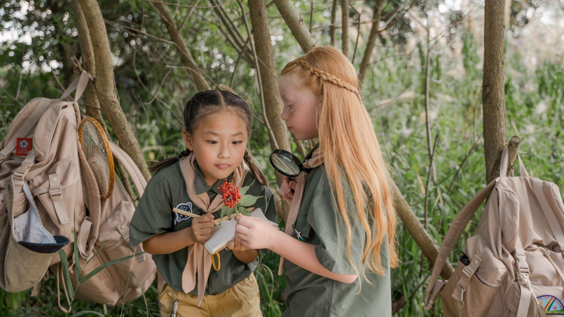 Two girls inspect  some berries with a magnifying glass while outsite.