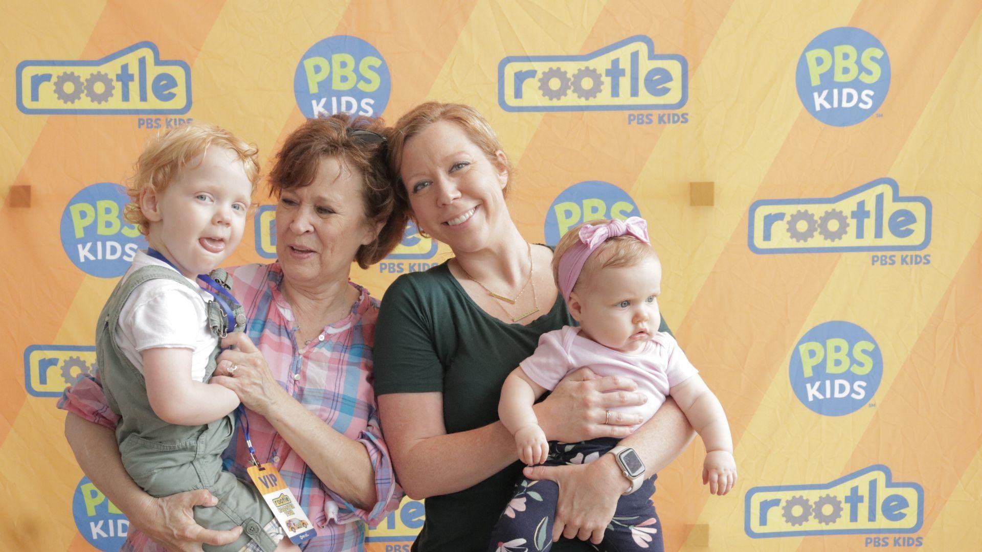 A family stands together in front of the Rootle PBS KIDS backdrop at a Rootle Roadster Tour stop.